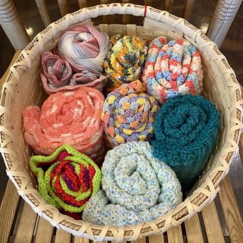 A basket sitting on a wooden rocking chair with seven rolled up, multicolored knitted afghans inside